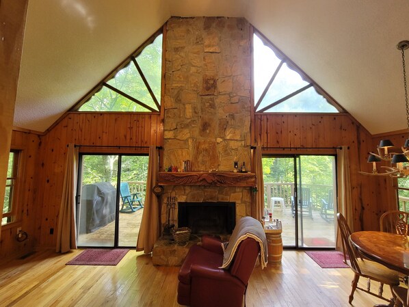 A cozy cabin interior with a stone fireplace, wooden walls, and large windows offering a view of the outdoors.