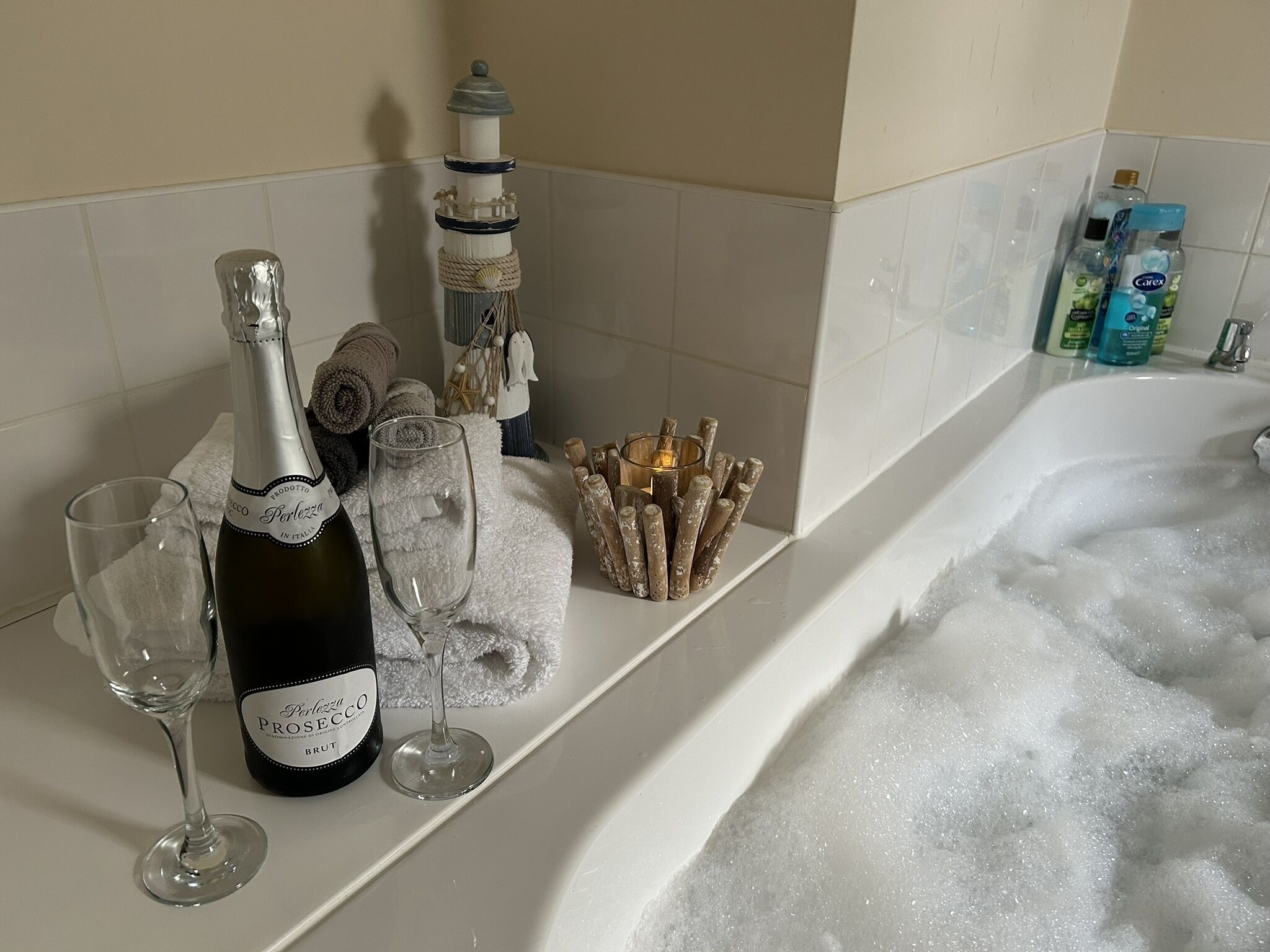 A bathroom with a bathtub filled with bubbles, a bottle of Prosecco, two wine glasses, and toiletries on the counter.