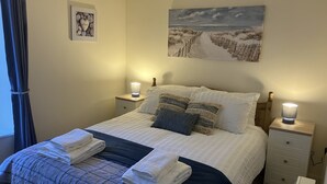 A neatly made bed with a white and dark blue quilt, two bedside lamps, a framed picture on the wall, and a painting above the bed. - Chapel St Apartments (Caernarfon)