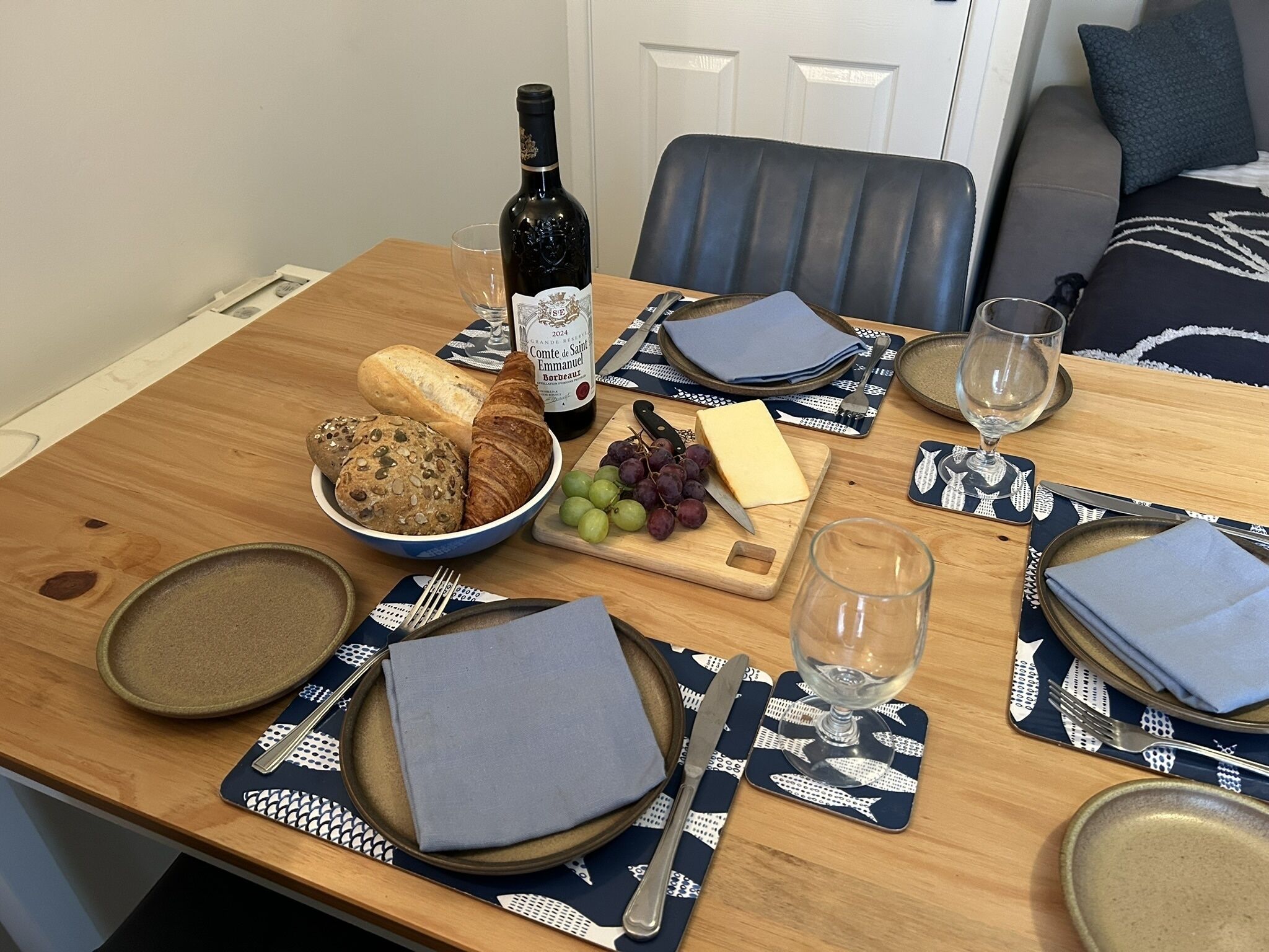 A dining table set with bread, cheese, grapes, a bottle of wine, and cutlery.