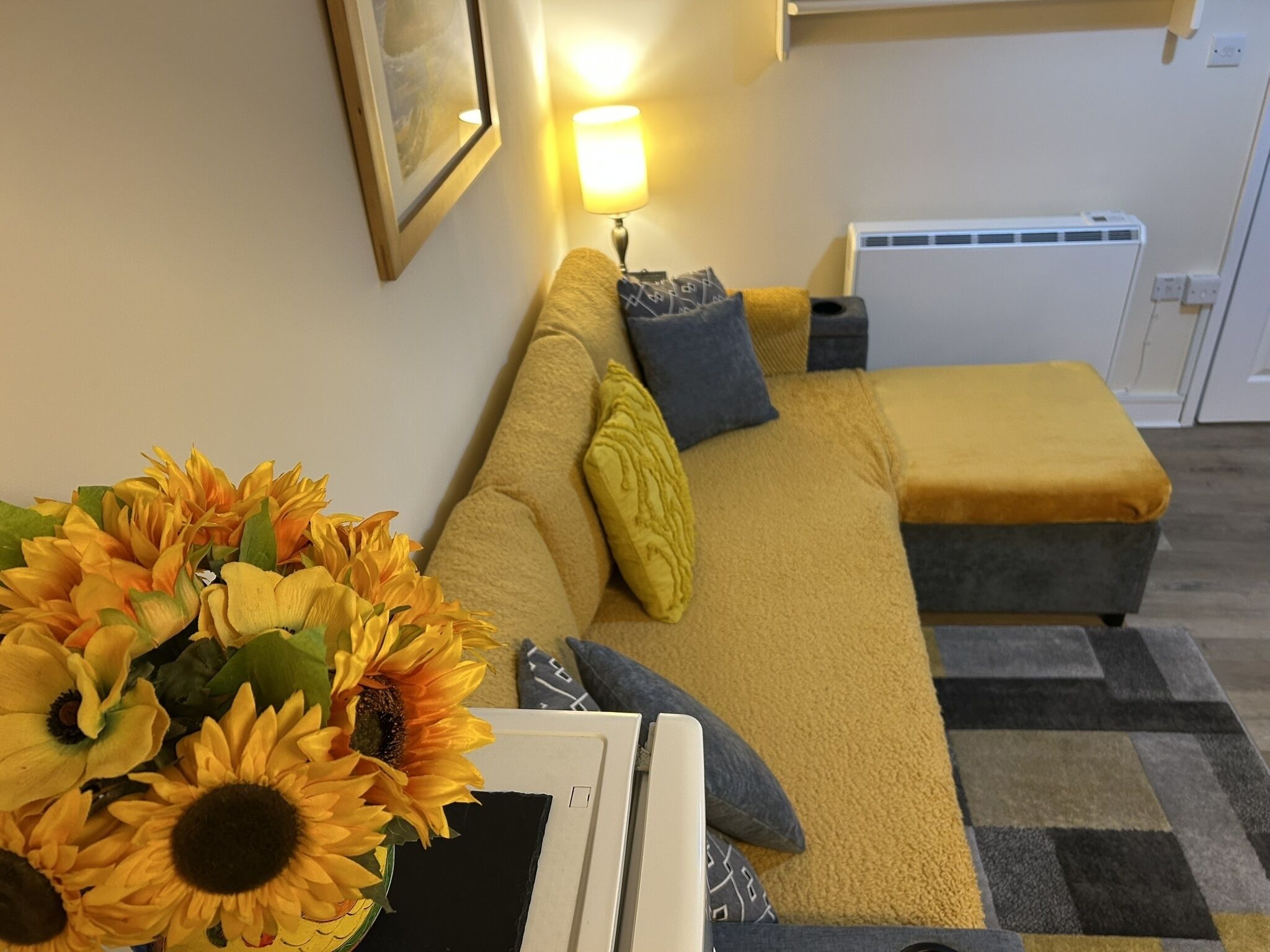 Ein gemütliches Wohnzimmer mit einer Couch, einem kleinen Tisch mit einer Vase voller Sonnenblumen, einer Lampe und einer Heizung.