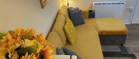 A cozy living room with a sofa, a small table with a vase of sunflowers, a lamp, and a radiator.