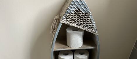 A corner shelf with a house-shaped design, holding toilet paper rolls and a small drawer.