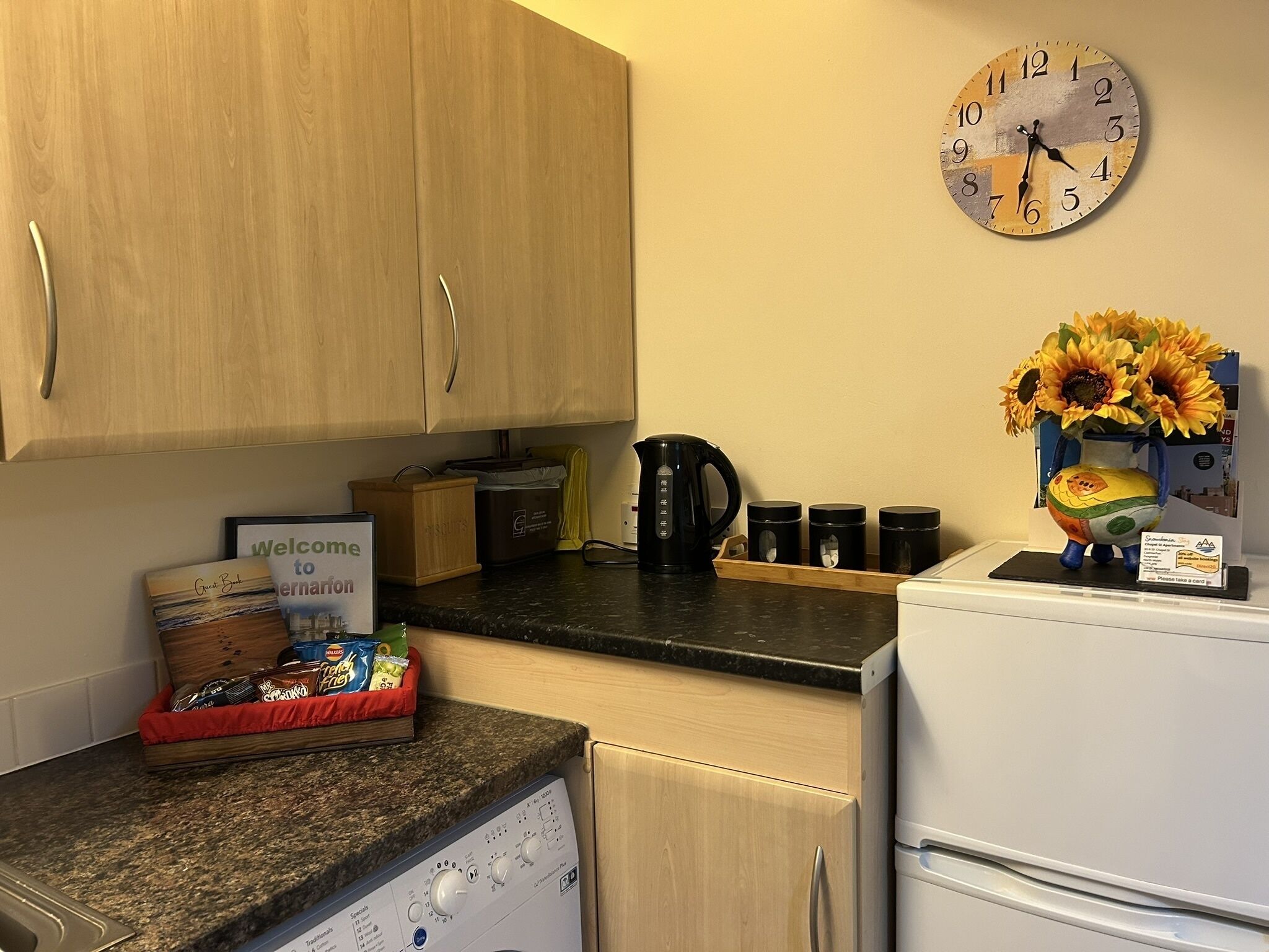 Een keuken met houten kastjes, een klok, een koffiezetapparaat en een vaas met zonnebloemen.