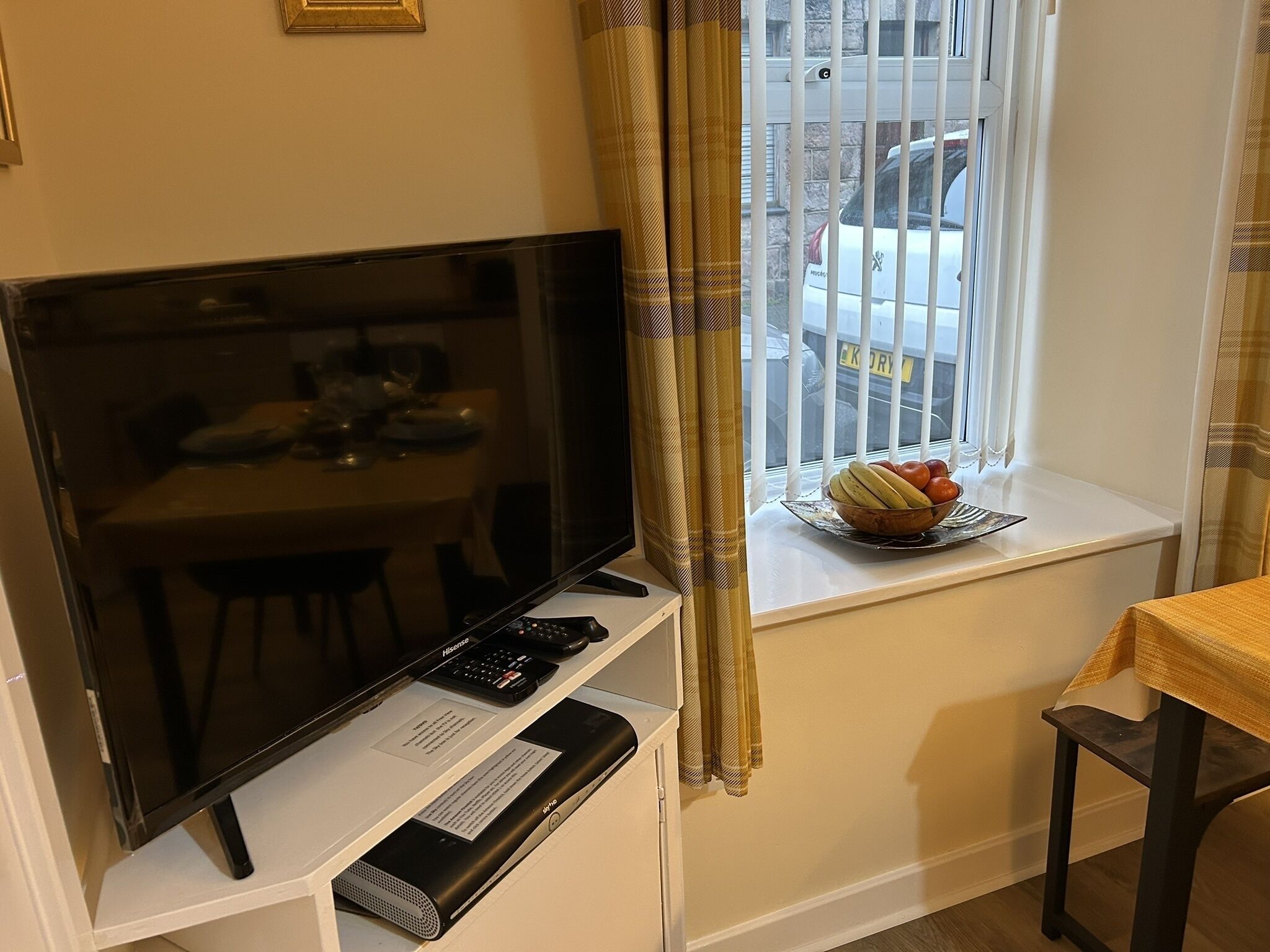 A room with a TV, a small table with a bowl of fruit, a window with a view of a parked car, and a curtain.