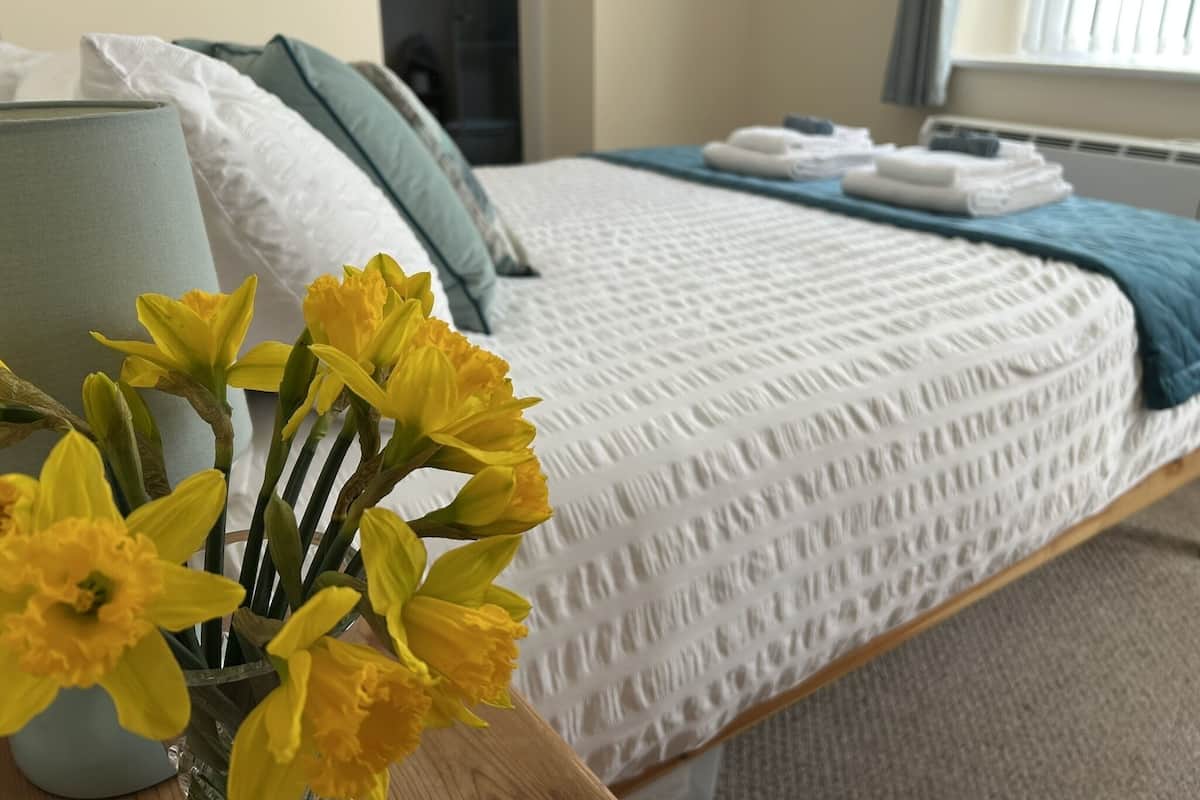 Un dormitorio con cama, almohadas, una mesita de noche con un jarrón de flores y una ventana con cortinas.