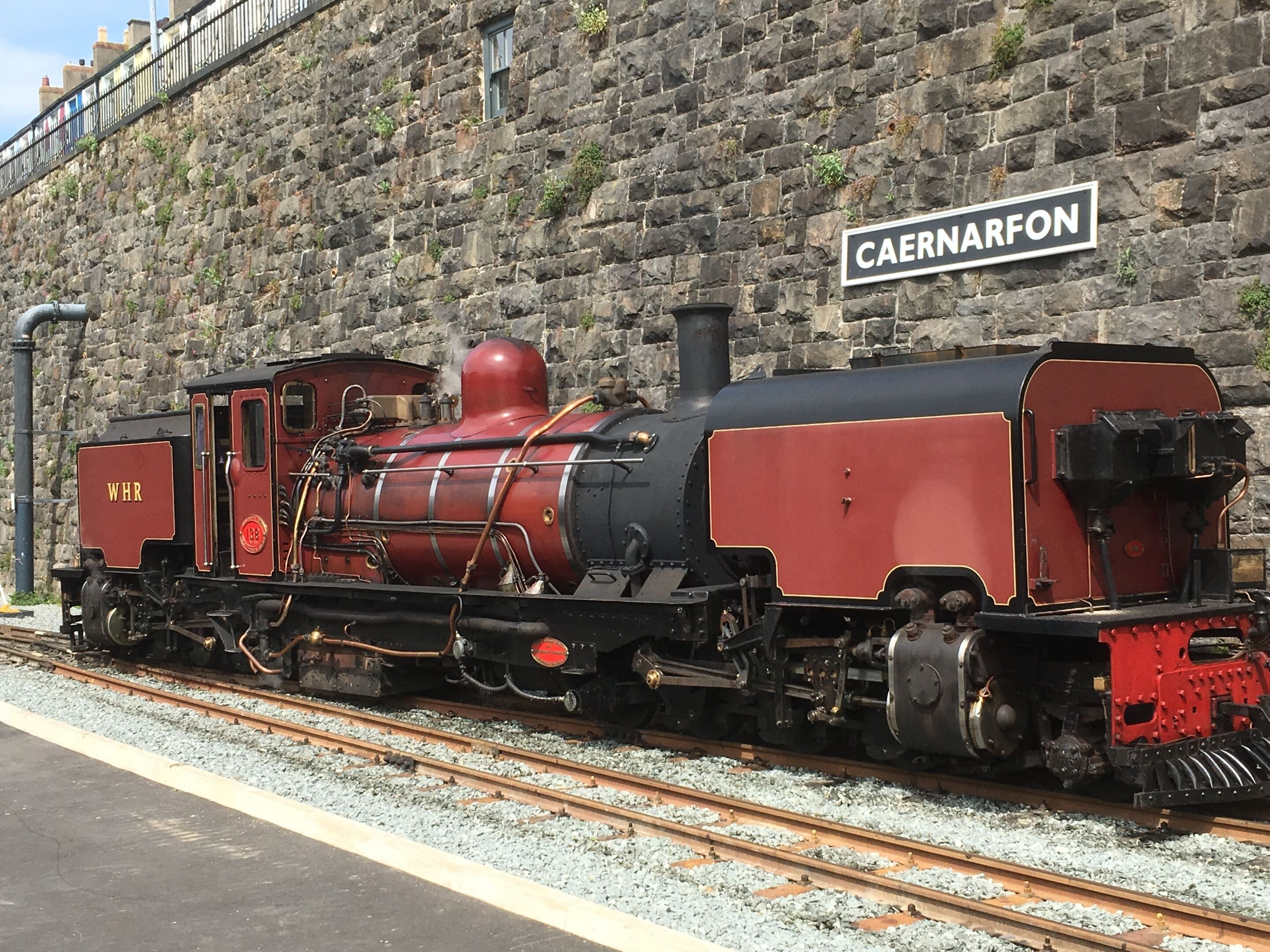 A vintage red and black steam locomotive at a railway station with a stone wall and a sign reading "CAERNARFON".