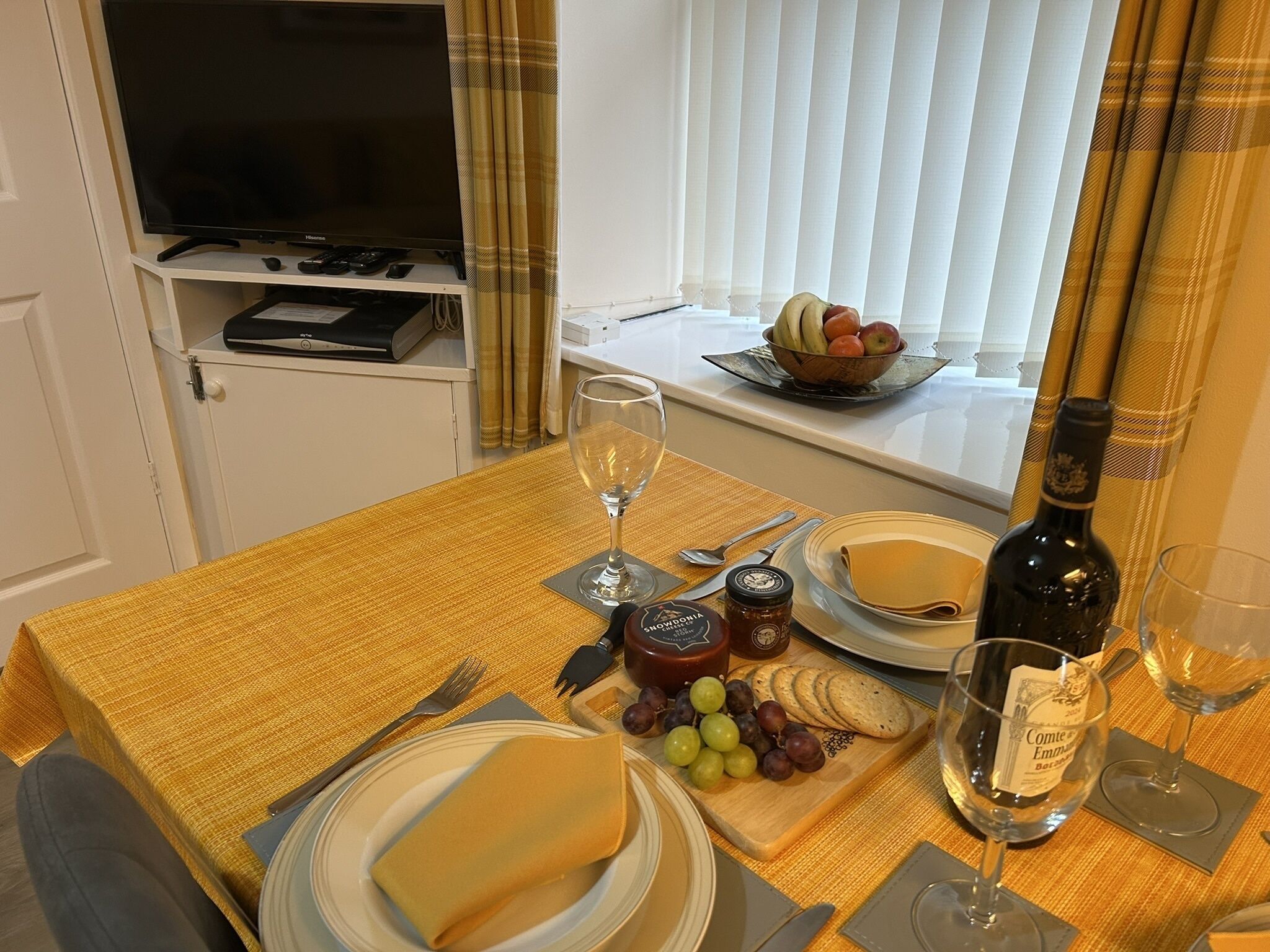 A dining table set with a bottle of wine, glasses, a bowl of fruit, and a plate of cheese.
