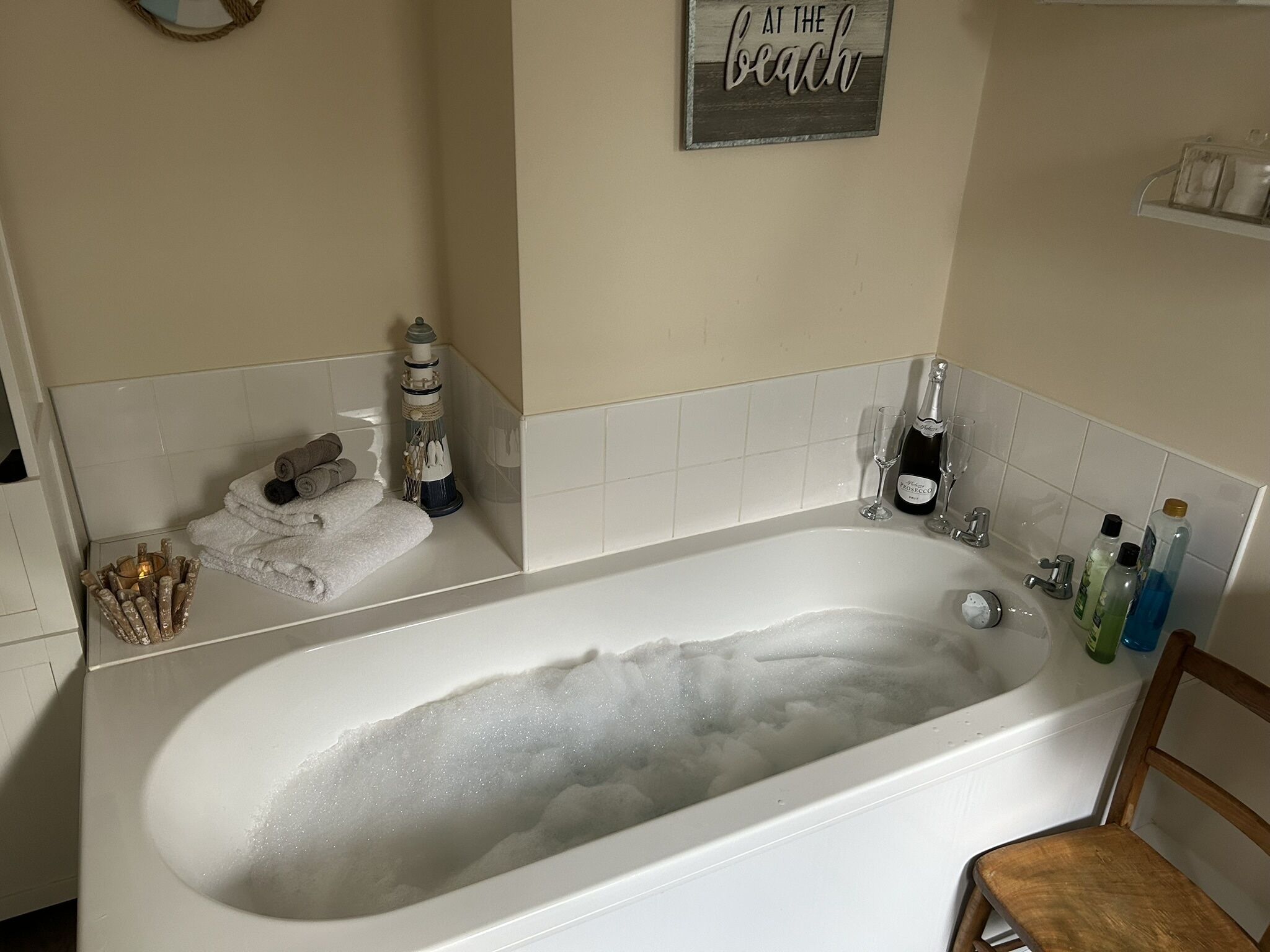 A bathroom with a large bathtub filled with bubbles, a towel rack with towels, a bottle of champagne, and a sign that says "At the beach".