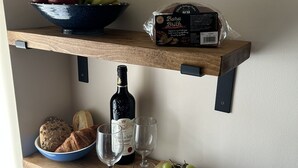 A kitchen shelf with a bowl of fruit, a loaf of bread, a bottle of wine, and a cutting board with cheese and grapes. - Chapel St Apartments (Caernarfon)