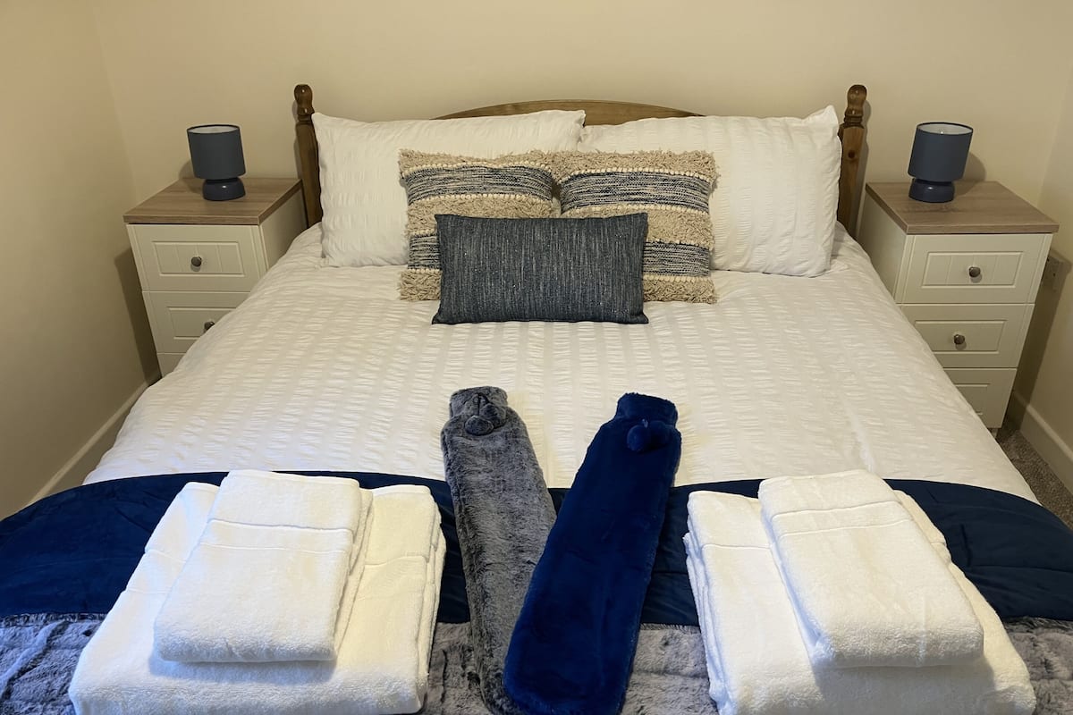 Una cama bien hecha con una colcha blanca, dos almohadas y una manta azul. Hay dos mesitas de noche con lámparas y un cajón pequeño.