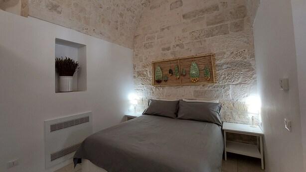 A bedroom with a stone wall, a bed with grey bedding, a bedside table with a lamp, and a small shelf with a plant.