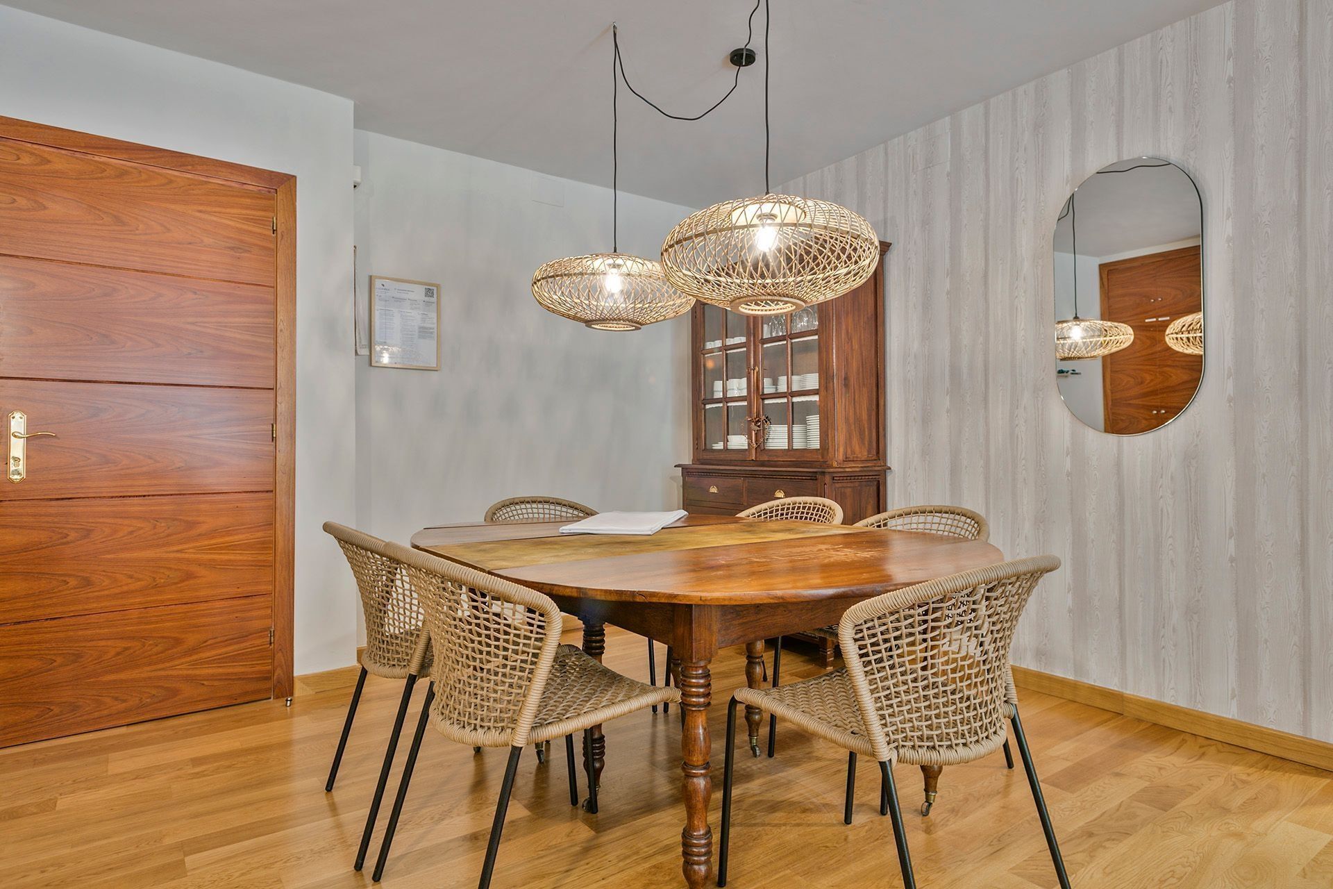 Un comedor con mesa de madera, sillas de mimbre, un armario de madera y un espejo en la pared.