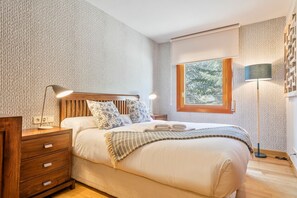 A bedroom with a wooden headboard, a bedside table with a lamp, a window with a view of trees, and a patterned wall.