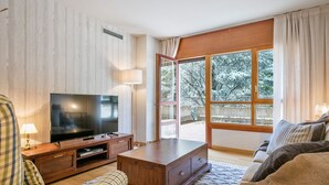 A living room with a TV, wooden furniture, a large window, and a view of trees.