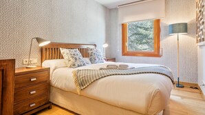 A bedroom with a wooden headboard, a bedside table with a lamp, a window with a view of trees, and a patterned wall.