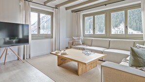 Ein modernes Wohnzimmer mit einem Couchtisch aus Holz, einer Couch und einem Fernseher auf einem Schrank. Große Fenster bieten Blick auf einen verschneiten Berg.