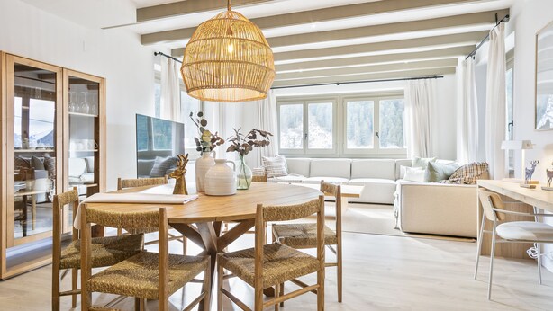 Ein moderner Essbereich mit einem Holztisch und Stühlen, einer großen gewebten Pendelleuchte und Blick auf eine verschneite Landschaft durch große Fenster.