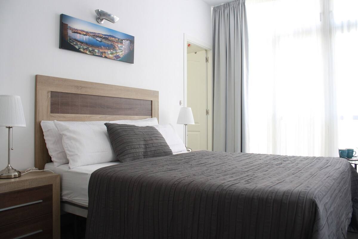 Una cama bien hecha con una manta gris, cabecero de madera y almohada blanca. Hay una mesita de noche con una lámpara y una taza. En la pared, encima de la cama, se ve un cuadro de un paisaje urbano.