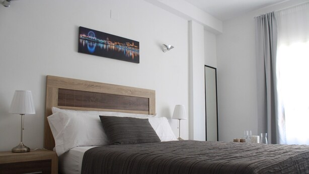 A hotel room with a wooden headboard, a large bed with dark bedding, two bedside lamps, a nightstand, and a wall-mounted picture of a cityscape. - The Catherdral Rooms (Málaga)