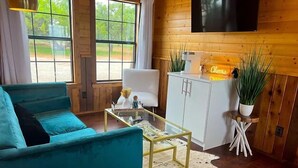 A cozy living room with a teal sofa, a glass coffee table, and a white cabinet. There are two potted plants on stands, and a cowhide rug on the floor.