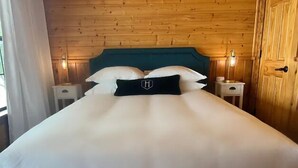 A neatly made bed with a teal headboard, white bedding, and a black pillow with a logo. There are two bedside tables with lamps, and a wooden door is visible to the right.