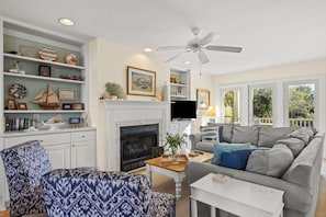 A living room with a fireplace, a ceiling fan, and a large window.
