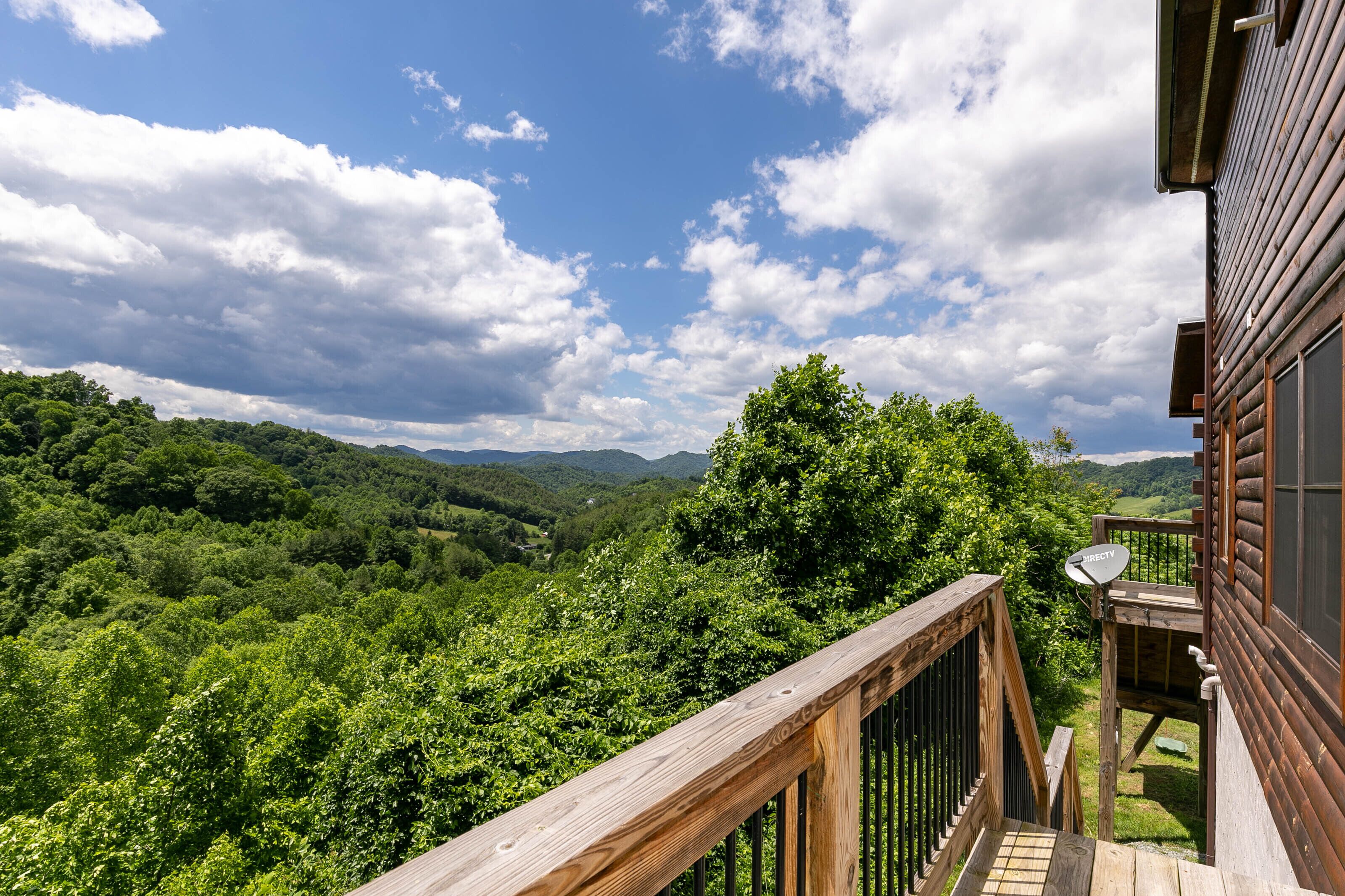A wooden deck with a satellite dish, overlooking a lush green valley and distant mountains under a partly cloudy sky.