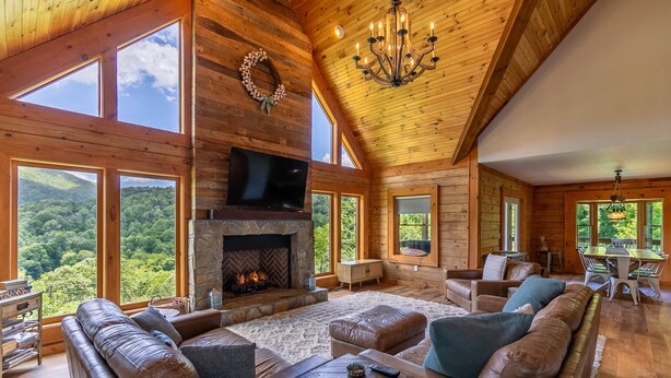 A spacious living room with a fireplace, leather sofas, a television, and large windows offering a view of the forest.