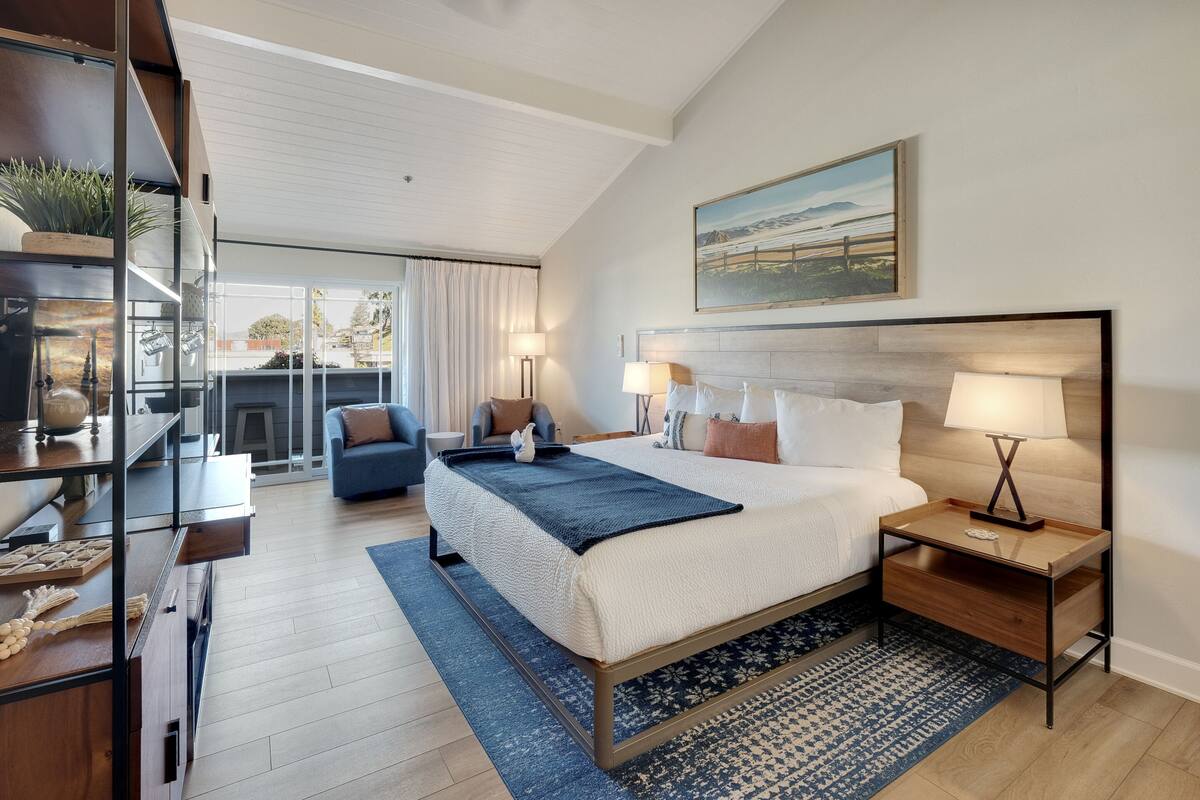 A modern bedroom with a large bed, bedside tables with lamps, a view of the outdoors, and a painting on the wall.