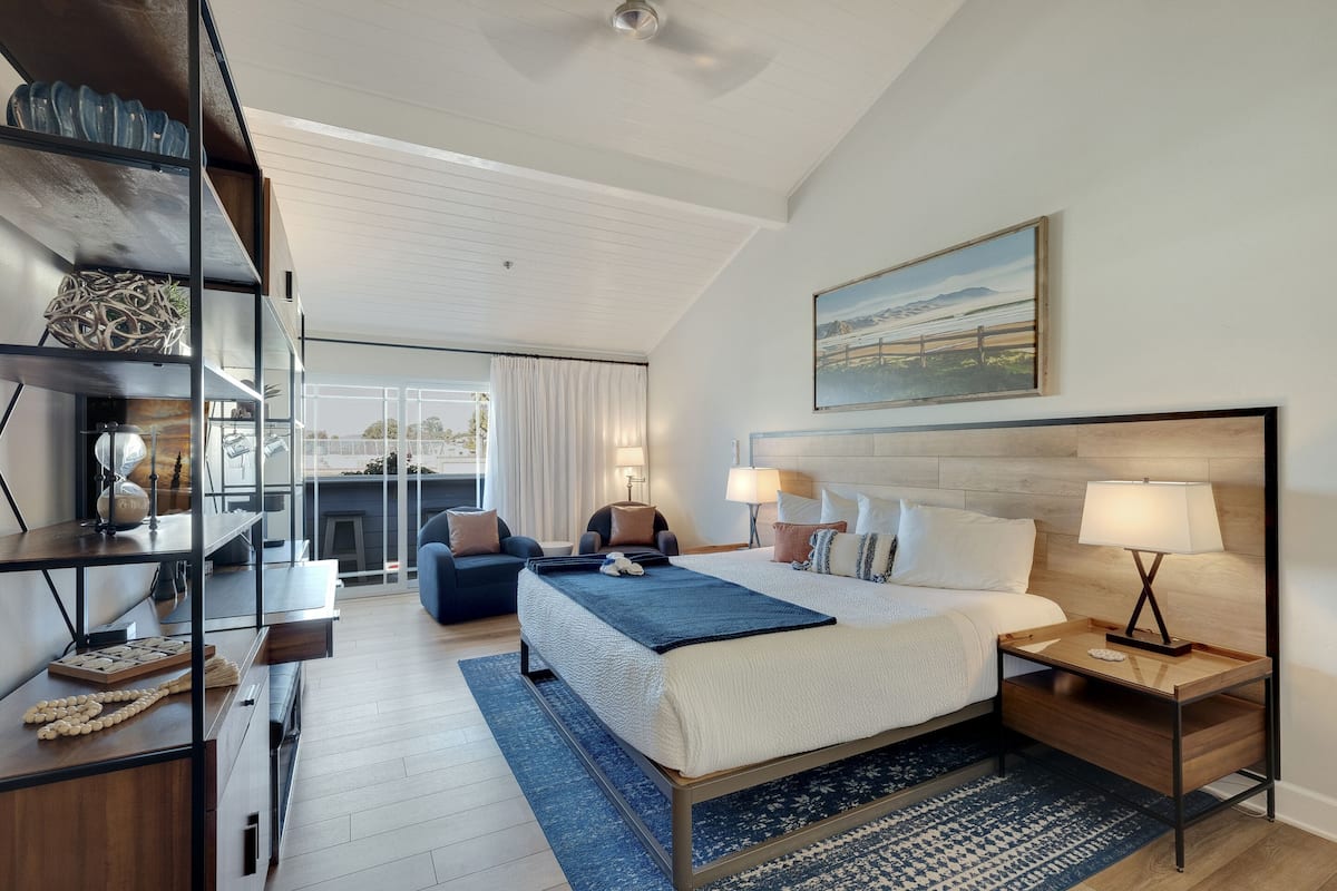 A modern hotel room with a large bed, bedside tables, a desk with a chair, a bookshelf, and a large window with a view.
