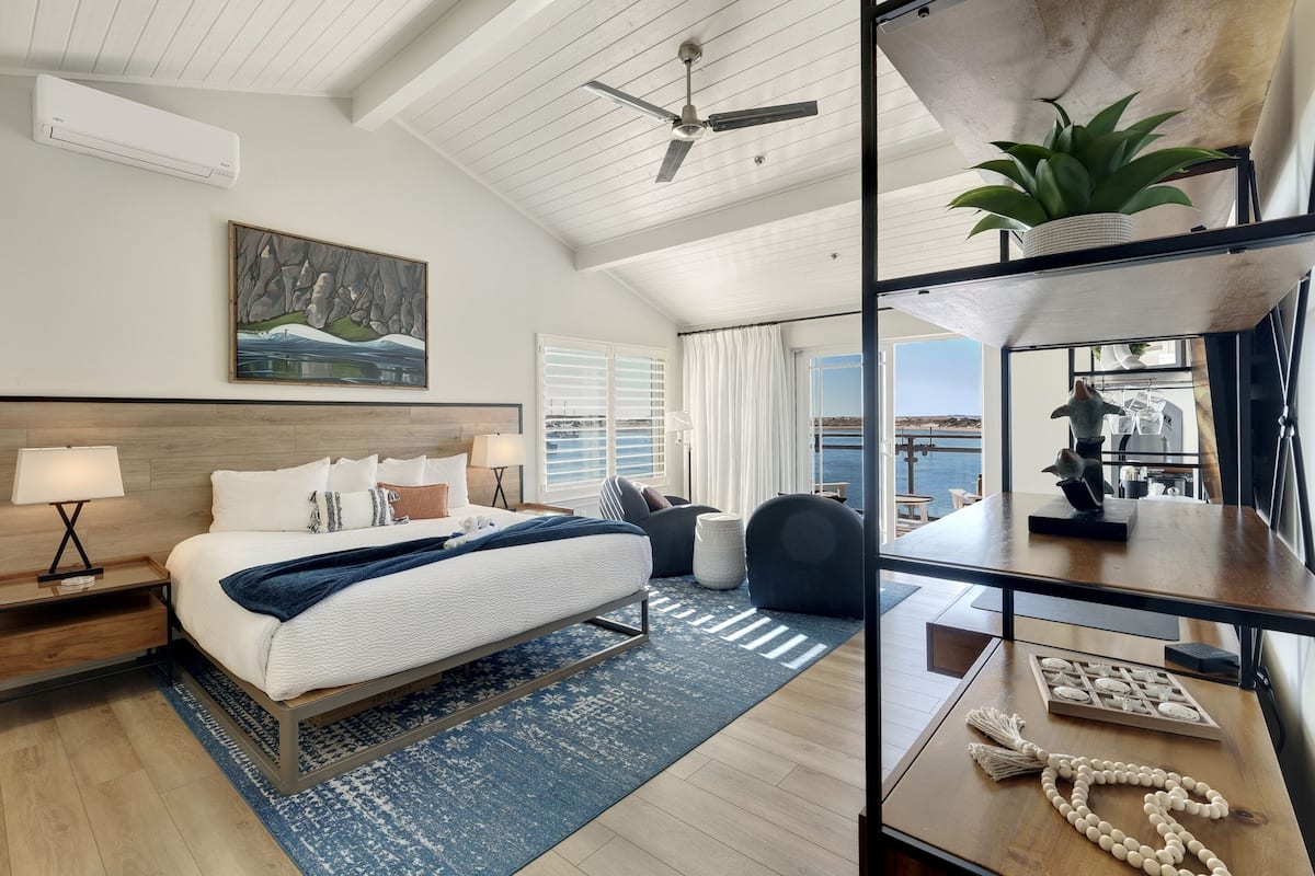 A modern bedroom with a large bed, a ceiling fan, a view of the sea, and a balcony with a potted plant.