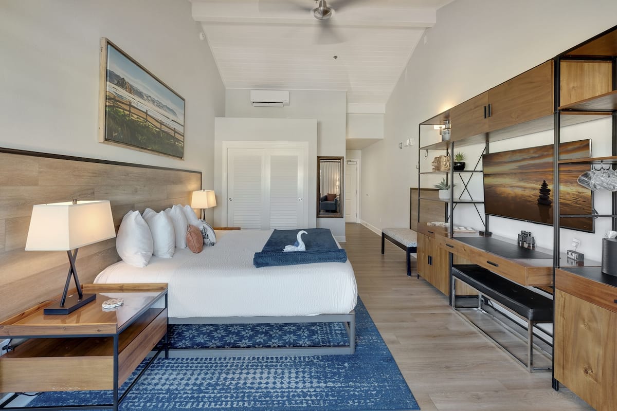 A modern hotel room with a large bed, a desk, a TV, and a bookshelf.