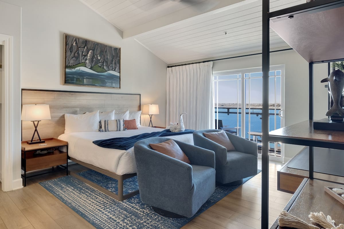 A hotel room with a large bed, two armchairs, a nightstand with a lamp, a painting on the wall, and a view of the sea through large windows.