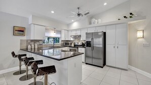 A modern kitchen with white cabinets, a stainless steel refrigerator, a central island with bar stools, and a ceiling fan.