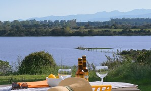 A lakeside table with a bottle, wine glass, and sunglasses, overlooking a calm lake with a dock and distant mountains. - 2-bedroom apartment on an 8-hectare estate, on the shores of Lake Teppe Rosse, 7 km from the beaches, air conditioning, Wi-Fi (Aléria)