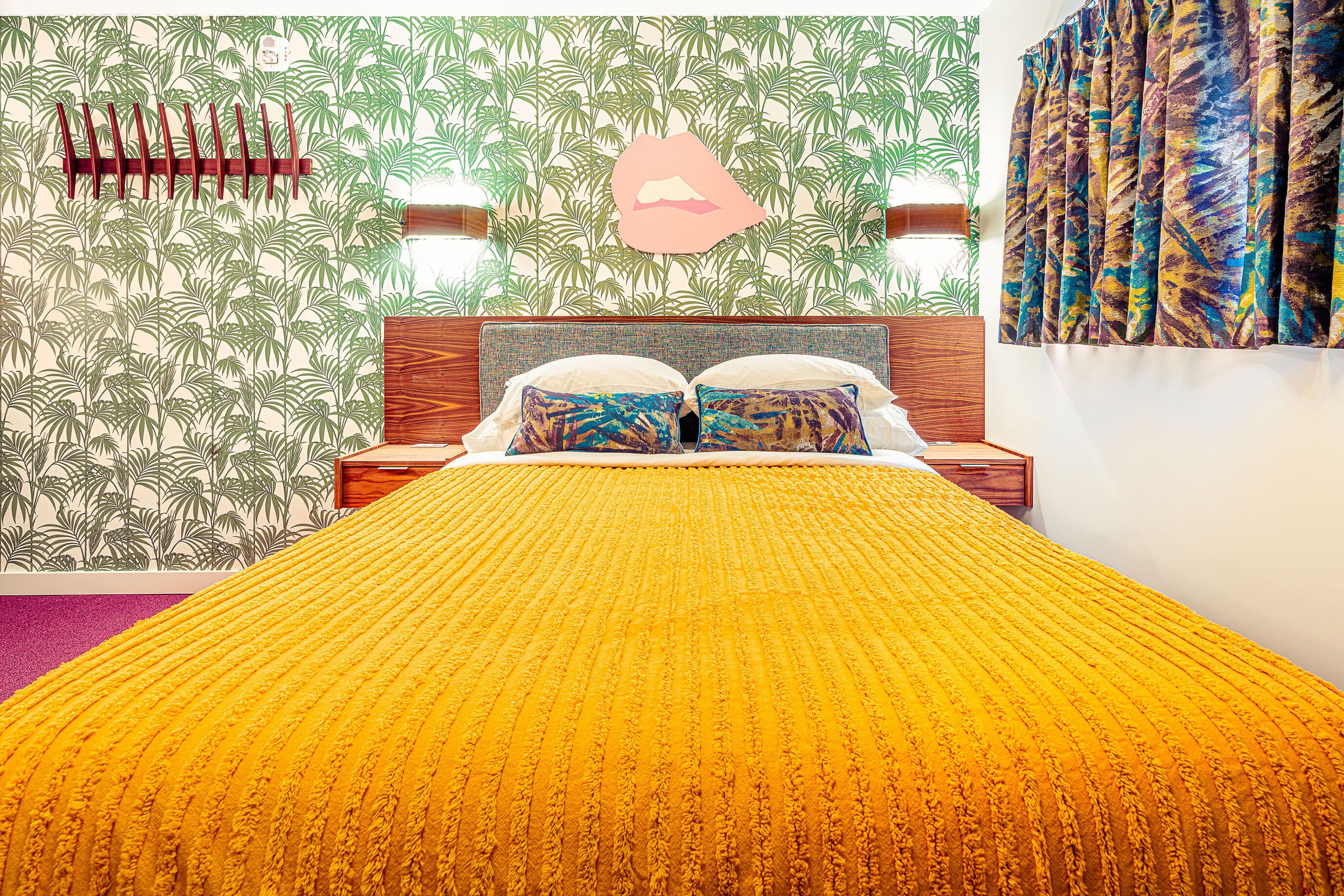 A bedroom with a bed, a headboard, a nightstand, a wall with a leaf pattern, and a curtain.