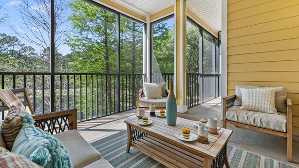 Un balcon ensoleillé avec un canapé, une table ornée d’un vase et une vue sur les arbres.