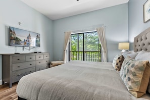 A bedroom with a large bed, a dresser, a television on the wall, and a balcony with curtains.
