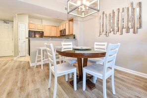 A dining area with a round table and white chairs, a kitchen with wooden cabinets, and a wall with wooden art pieces. - Resort Condo w/ Lagoon-Style Pool Views in Central Destin! ☀🌴🏖 (Destin)
