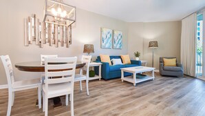 A living room with a blue sofa, white chairs, a coffee table, and a dining table. There are abstract paintings on the wall and a floor lamp. - Resort Condo w/ Lagoon-Style Pool Views in Central Destin! ☀🌴🏖 (Destin)