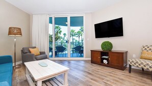 A living room with a sofa, a coffee table, a TV mounted on the wall, and a view of palm trees through the glass doors. - Resort Condo w/ Lagoon-Style Pool Views in Central Destin! ☀🌴🏖 (Destin)