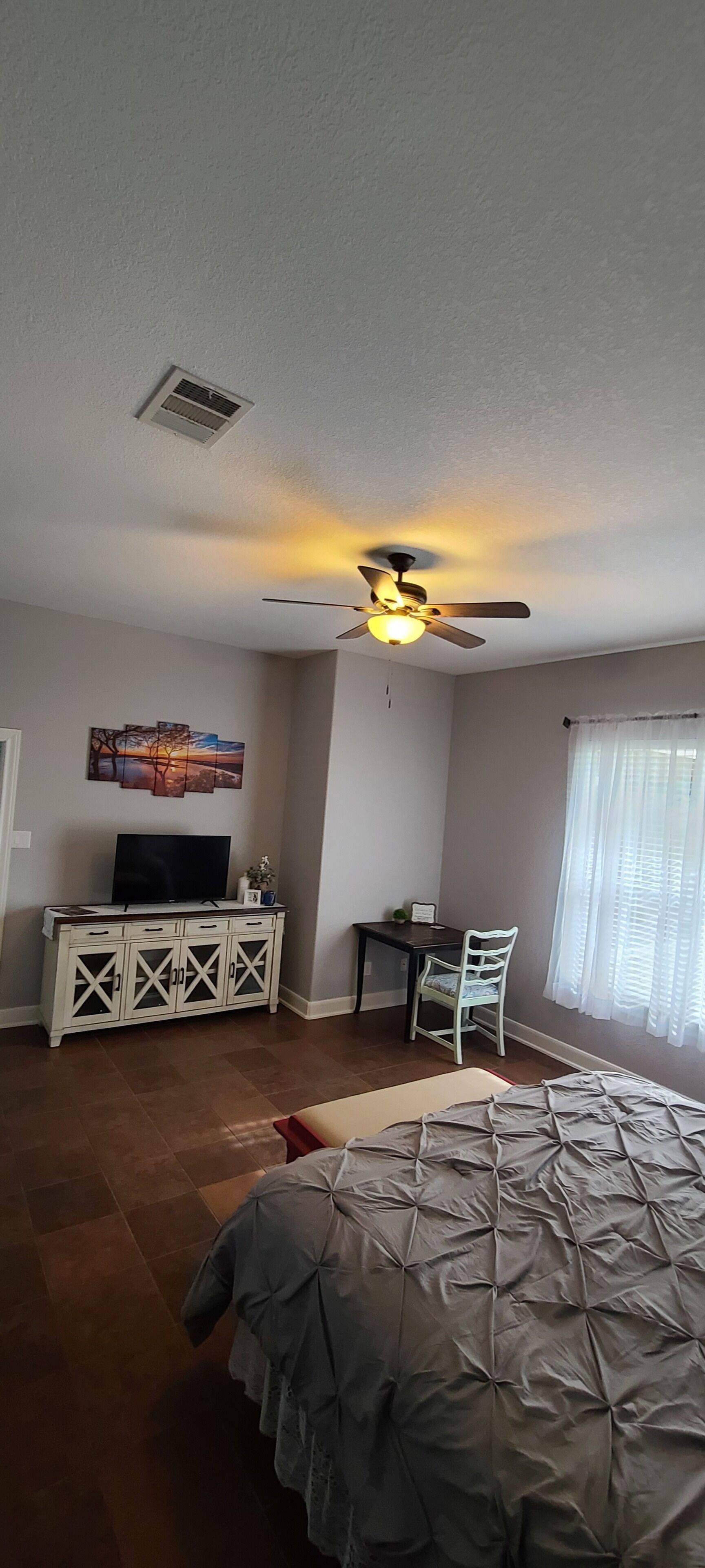 A bedroom with a bed, a ceiling fan, a TV on a cabinet, a desk with a chair, and a window with curtains.
