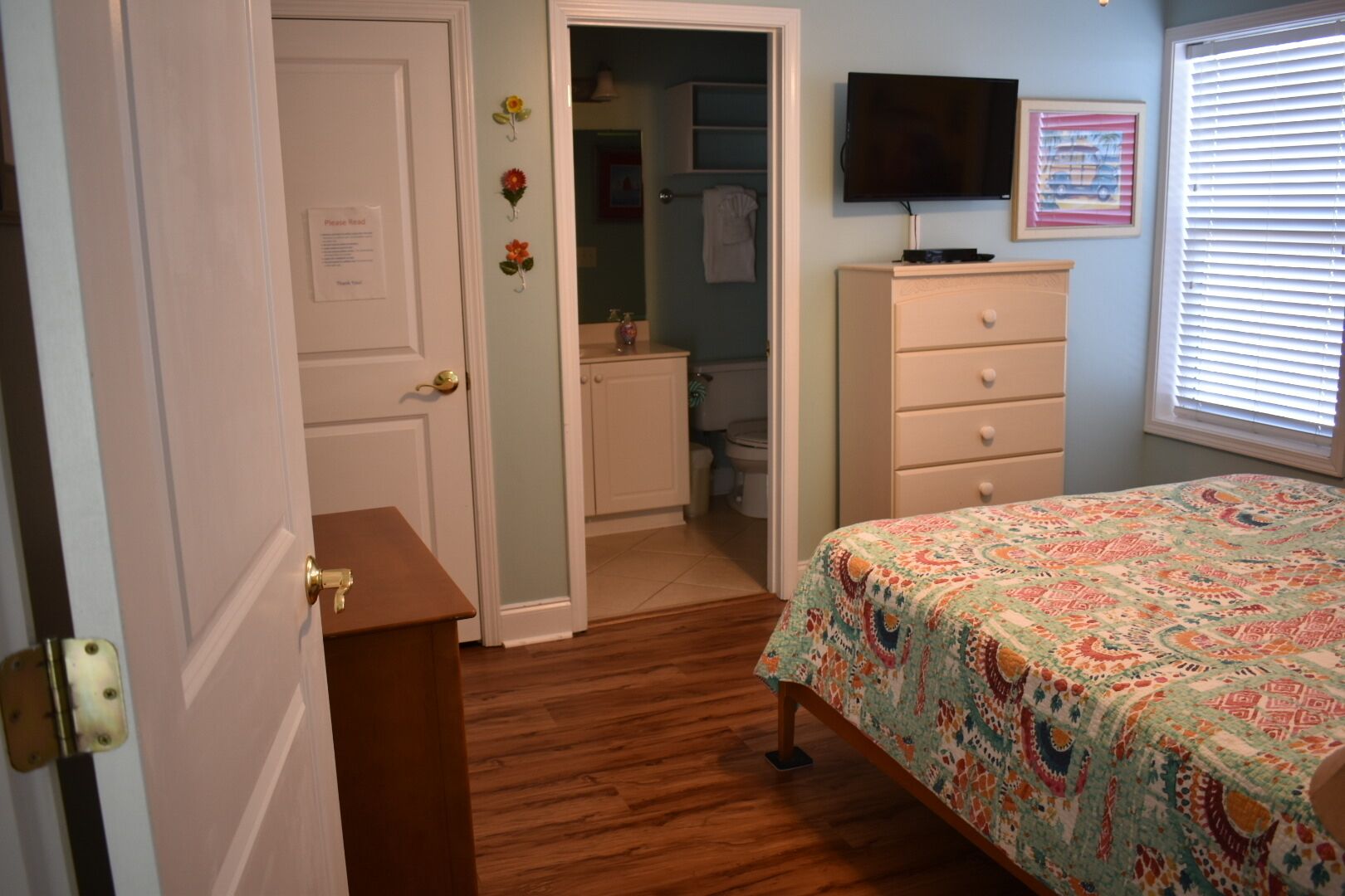 Une chambre avec un plancher en bois, un lit recouvert d’une couvre-lit à motifs, une commode, un téléviseur fixé au mur et une porte donnant sur une salle de bain.