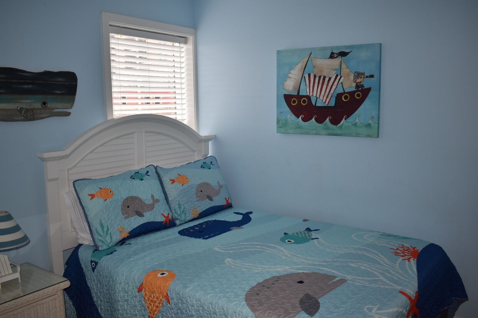 A bedroom with a bed featuring a nautical-themed bedspread, a white headboard, and a window with blinds.
