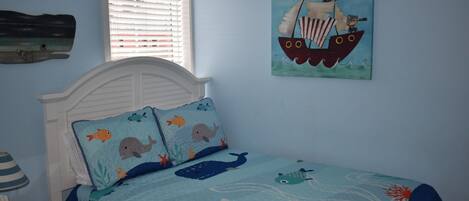 A bedroom with a bed featuring a nautical-themed bedspread, a white headboard, and a window with blinds.