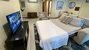 Quarto de hotel com uma cama grande, duas poltronas, uma TV em um suporte e uma mesinha.