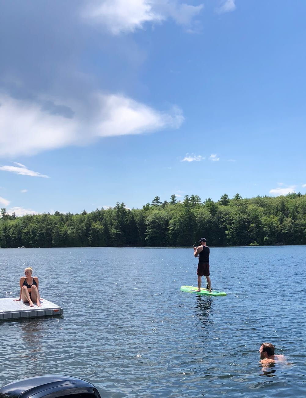 A lake with a dock, a person standing on a green paddleboard, and another person sitting on the dock.
