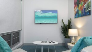 A room with a television displaying a beach scene, a round table with a magazine, two remote controls, a lamp, and teal cushions on white sofas. - Suite 1 with Kitchenette (St. John)