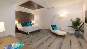 A modern hotel room with a bed, a desk, a chair, a sofa, a potted plant, and a wall-mounted lamp. - Suite 2, 10% Off Fall Stays (Cruz Bay)